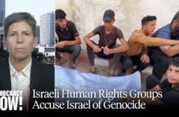 Israeli human rights activists holding banners and speaking at a press conference, accusing their own government of genocide in Gaza; expressions are solemn and serious, with signs calling for justice and an end to the violence.