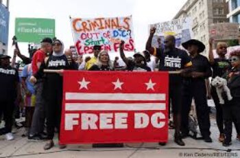 Protesters in Washington, D.C., holding signs and chanting in the streets at night, facing a line of National Guard troops in uniform and riot gear after President Trump ordered a federal takeover of the city’s police force and deployed military personnel.